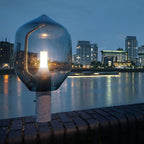 A Clérachic table lamp on white marble, lit at dusk on a river wall with a city skyline glowing behind it.
