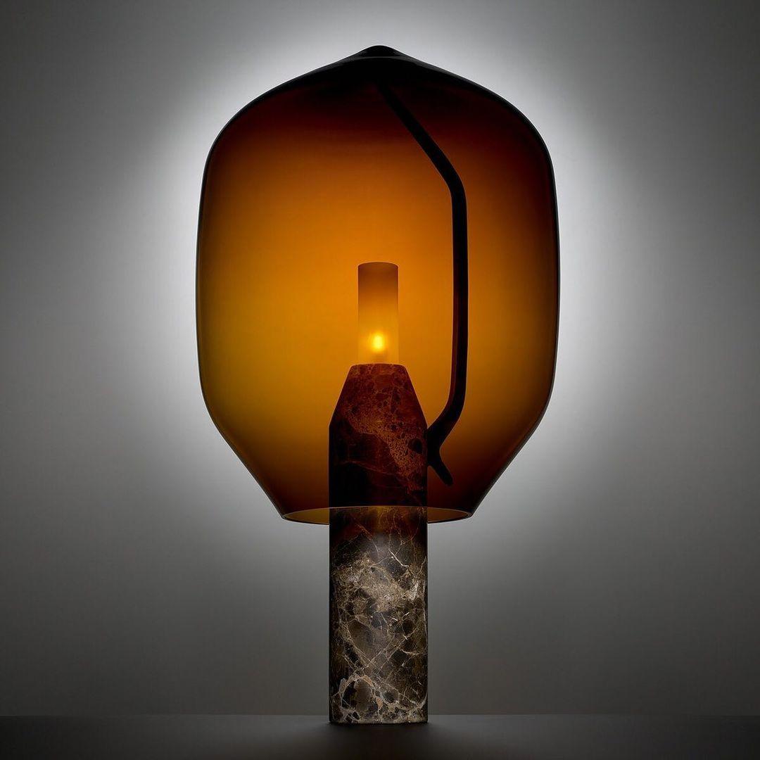 Clérachic's amber-glass lamp on a dark veined marble base, backlit against a charcoal background from another angle.