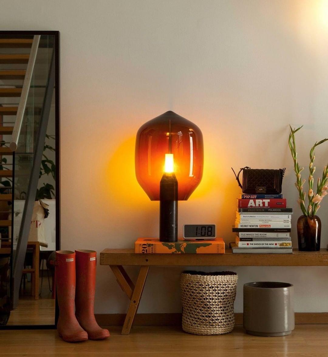 Clérachic's amber-glass table lamp lit in a hallway, beside red rain boots, woven baskets and a stack of books.