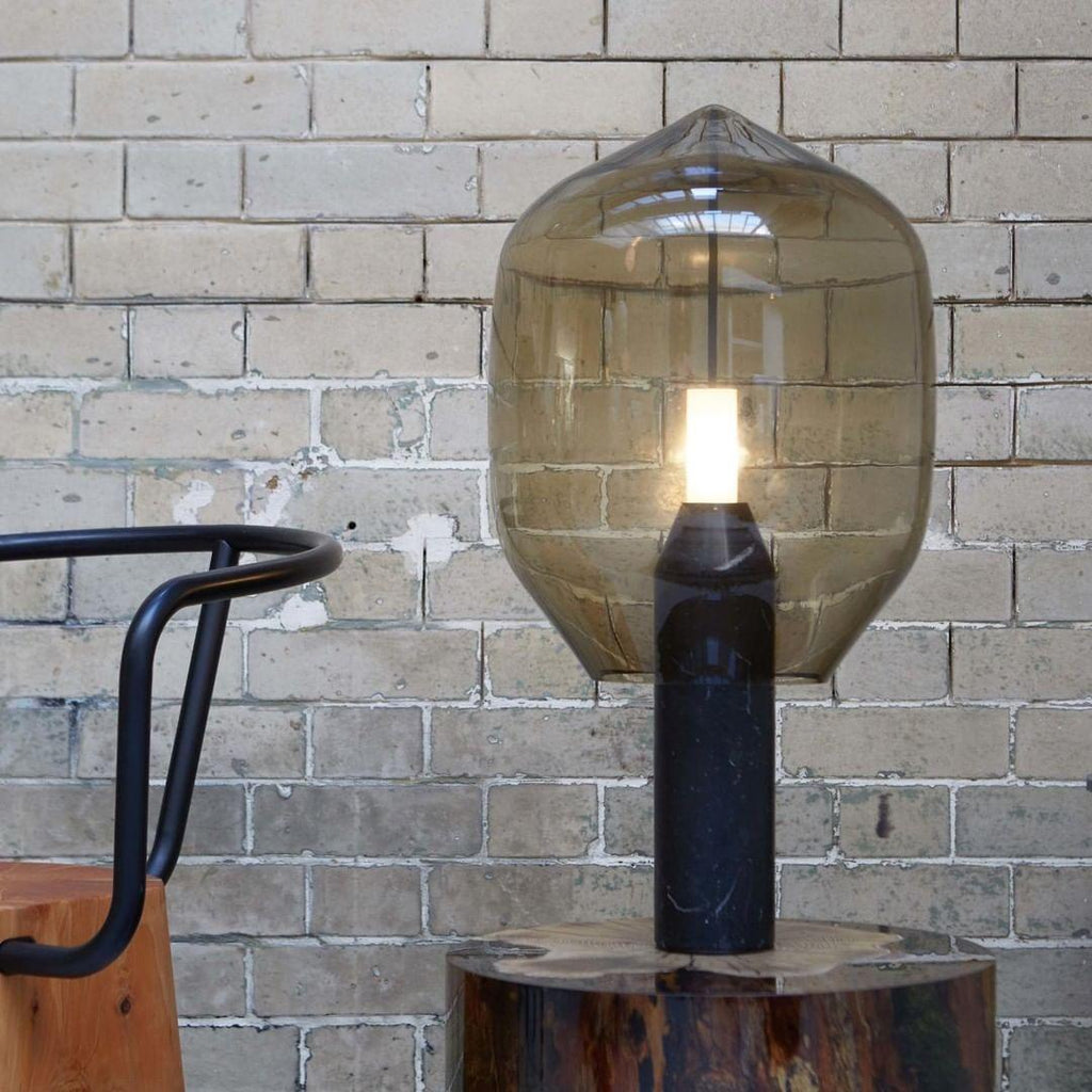 A smoked-glass Clérachic lamp on its black base, standing on a log slab against a weathered grey brick wall.