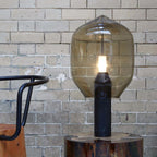 A smoked-glass Clérachic lamp on its black base, standing on a log slab against a weathered grey brick wall.