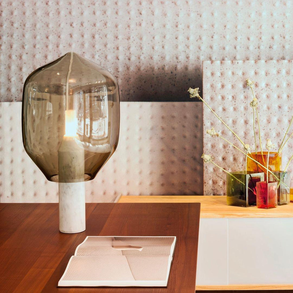 Clérachic's olive-toned glass lamp on a white marble base, set on a wood desk beside dried flowers and patterned wallpaper.