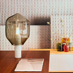 Clérachic's olive-toned glass lamp on a white marble base, set on a wood desk beside dried flowers and patterned wallpaper.
