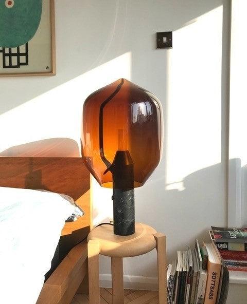 An amber-glass Clérachic lamp on a dark stone base, lit on a wood stool beside a sunlit bed and a stack of books.