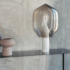 Clérachic's smoked-glass lamp on a white marble base, set on a shelf against a bare concrete wall next to a pink vase.