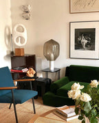 Clérachic's smoked-glass table lamp on white marble, standing in a living room beside a green velvet sofa and white roses.