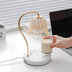 A hand places a glass candle jar beneath this Clérachic candle warmer lamp's marble base and brass gooseneck arm.