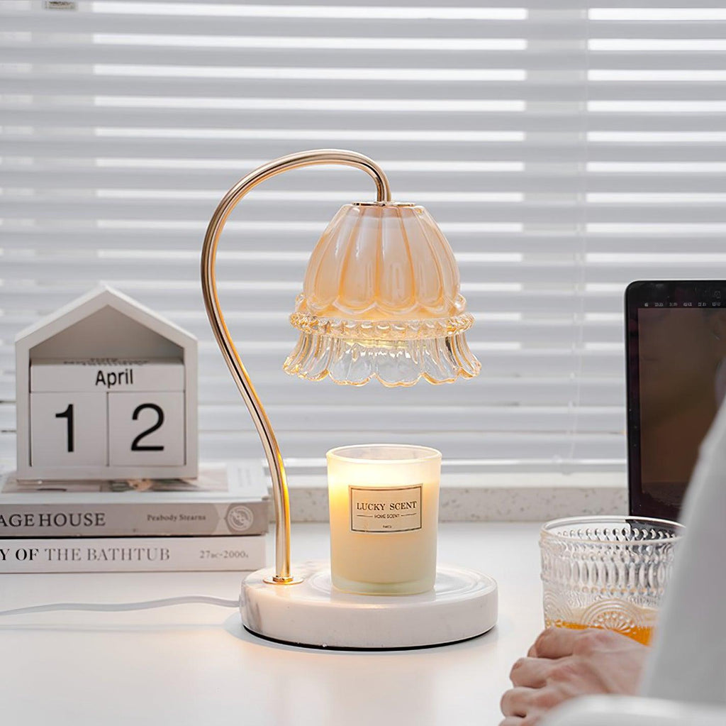 A Clérachic candle warmer lamp sits on a desk beside venetian blinds, with a hand reaching for a drink glass near its candle jar.