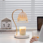 A Clérachic candle warmer lamp sits on a desk beside venetian blinds, with a hand reaching for a drink glass near its candle jar.