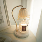 On a white side table beside a framed print, this Clérachic candle warmer lamp glows warm above its candle in a glass jar.