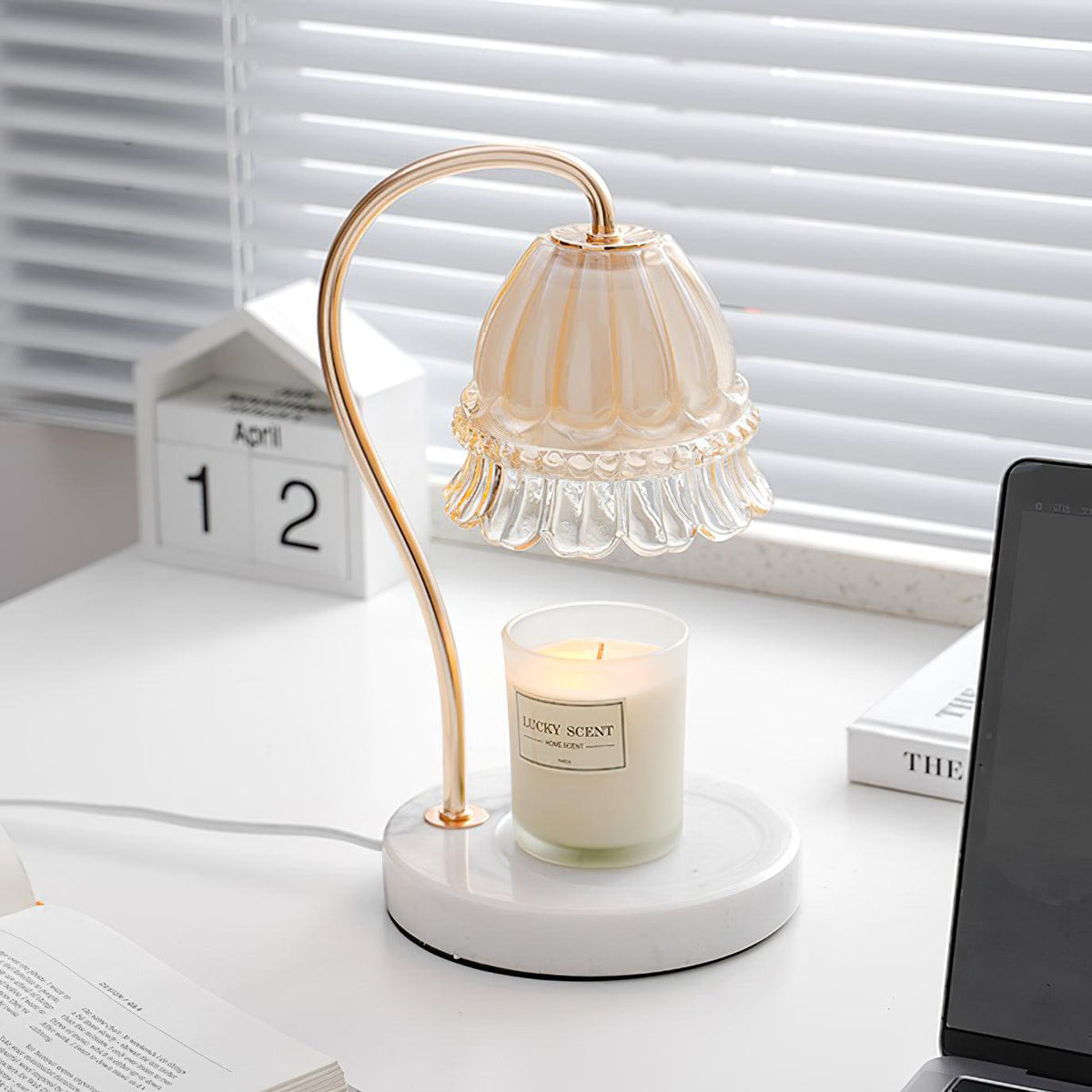 Beside a wooden desk calendar and venetian blinds, this Clérachic candle warmer lamp's amber glass shade glows over its candle.