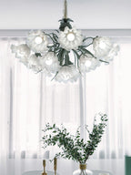 Framed by sheer white curtains, this Clérachic chandelier's shades catch the light, unlit, beside a vase of greenery.