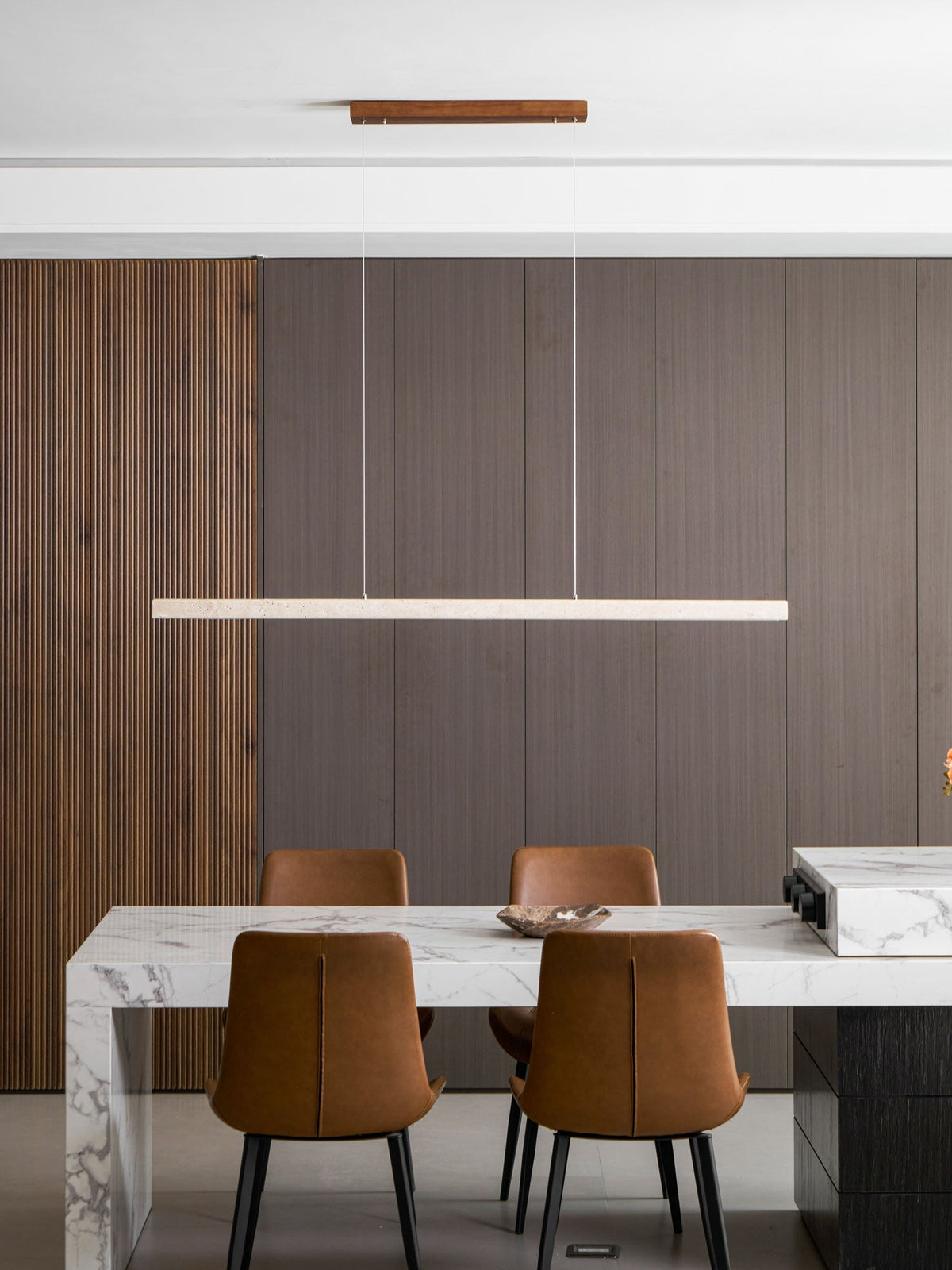 This Clérachic pendant hangs low above a marble dining table, its travertine bar glowing warm against ribbed wood panelling.