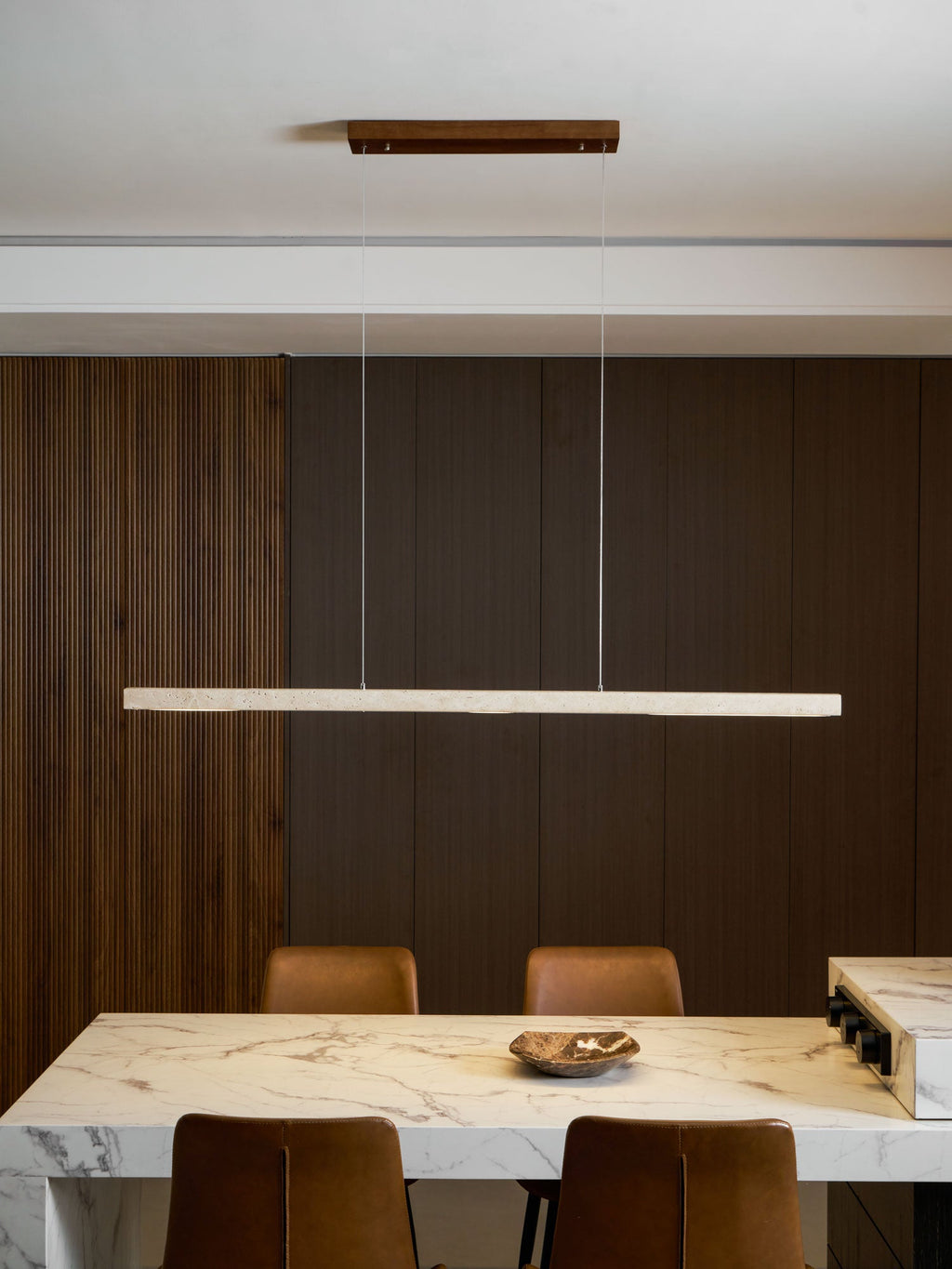 In a dining room with dark wood panelling, this Clérachic pendant's travertine bar glows warm above leather chairs and a marble table.