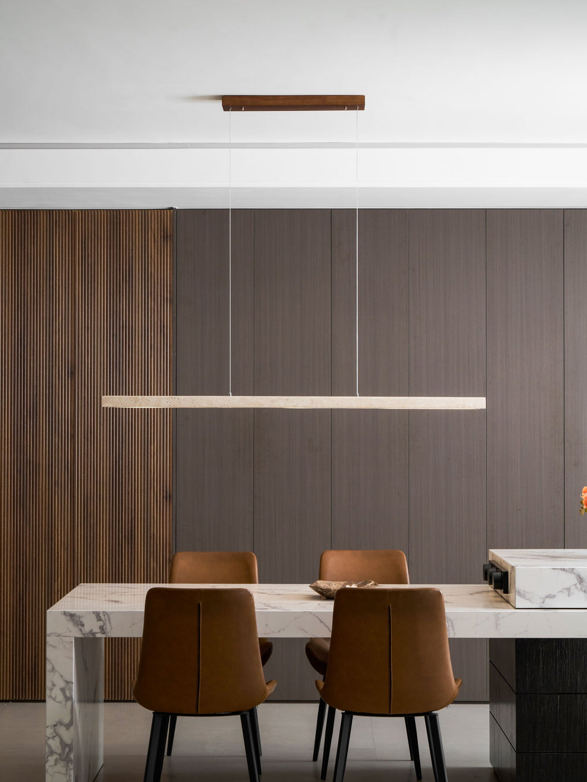 This Clérachic pendant hangs from a pale wood canopy, its travertine bar glowing warm above a marble dining table.