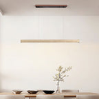 In a minimalist dining room, this Clérachic pendant's travertine bar glows warm above a table set with bowls and a bare branch.