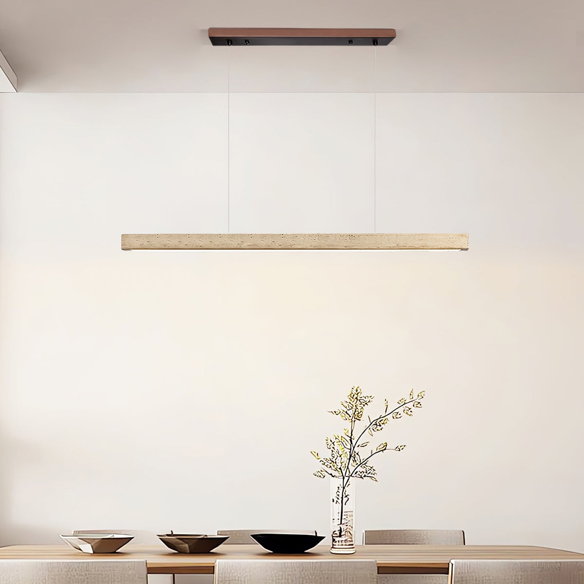 In a minimalist dining room, this Clérachic pendant's travertine bar glows warm above a table set with bowls and a bare branch.