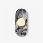 This Clérachic wall sconce shows a capsule-shaped natural stone plaque with a single warm glass orb centred on its veined grey surface, on a plain backdrop.