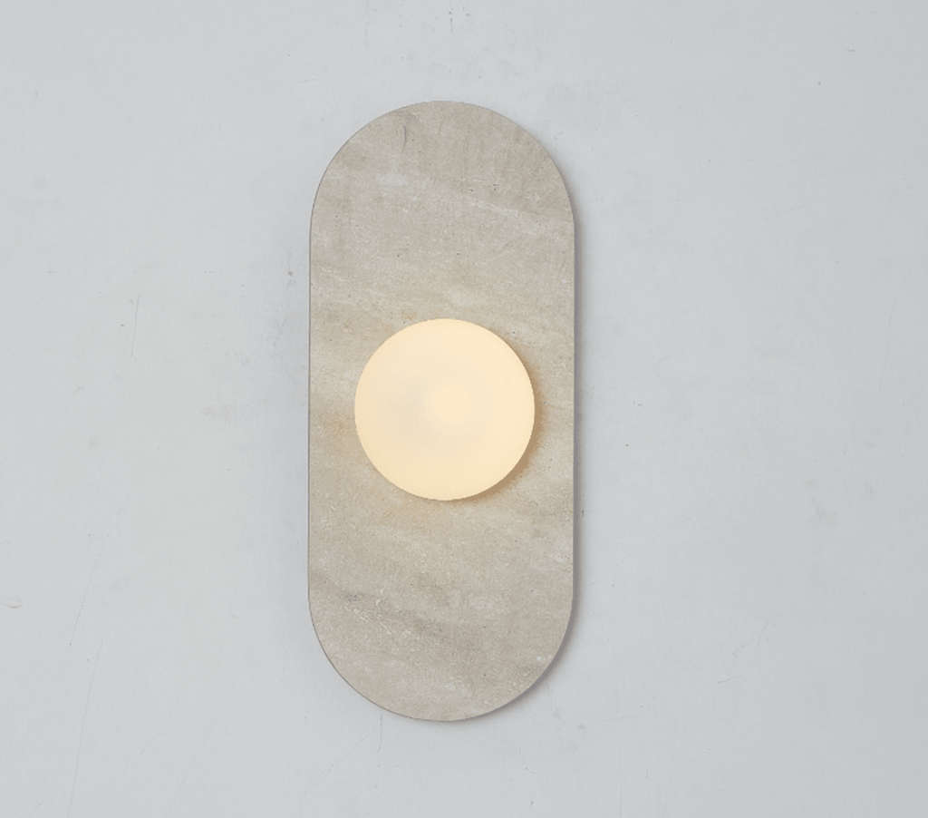 A beige travertine Clérachic wall sconce glows alone on a plain wall, its round glass globe lit warm at its centre.