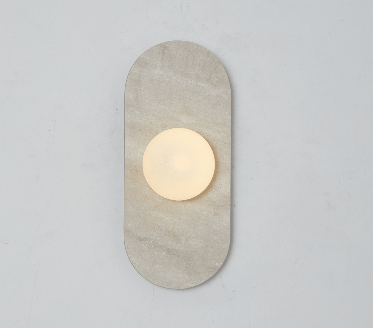 A beige travertine Clérachic wall sconce glows alone on a plain wall, its round glass globe lit warm at its centre.