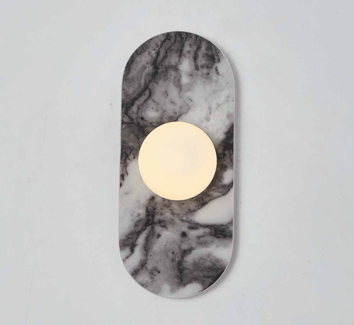 A pale grey marble Clérachic wall sconce hangs isolated against a plain wall, its glass globe glowing softly at its centre.