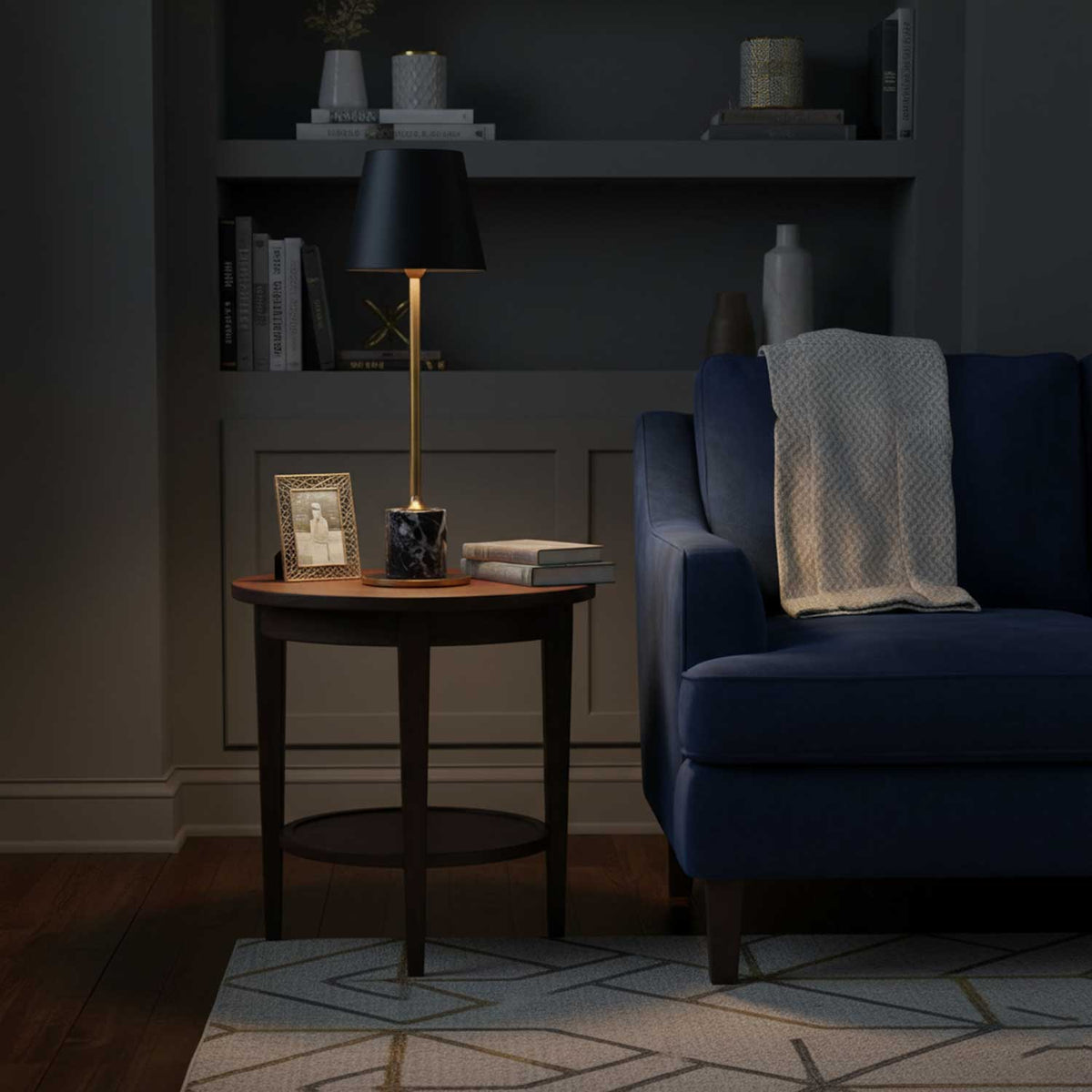 A Clérachic marble table lamp lights a round side table next to a navy armchair, books and a framed photo nearby on the shelf.