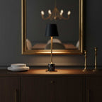 A Clérachic marble table lamp stands on a dark sideboard below a gilt-framed mirror, brass candlesticks and stacked plates beside it.