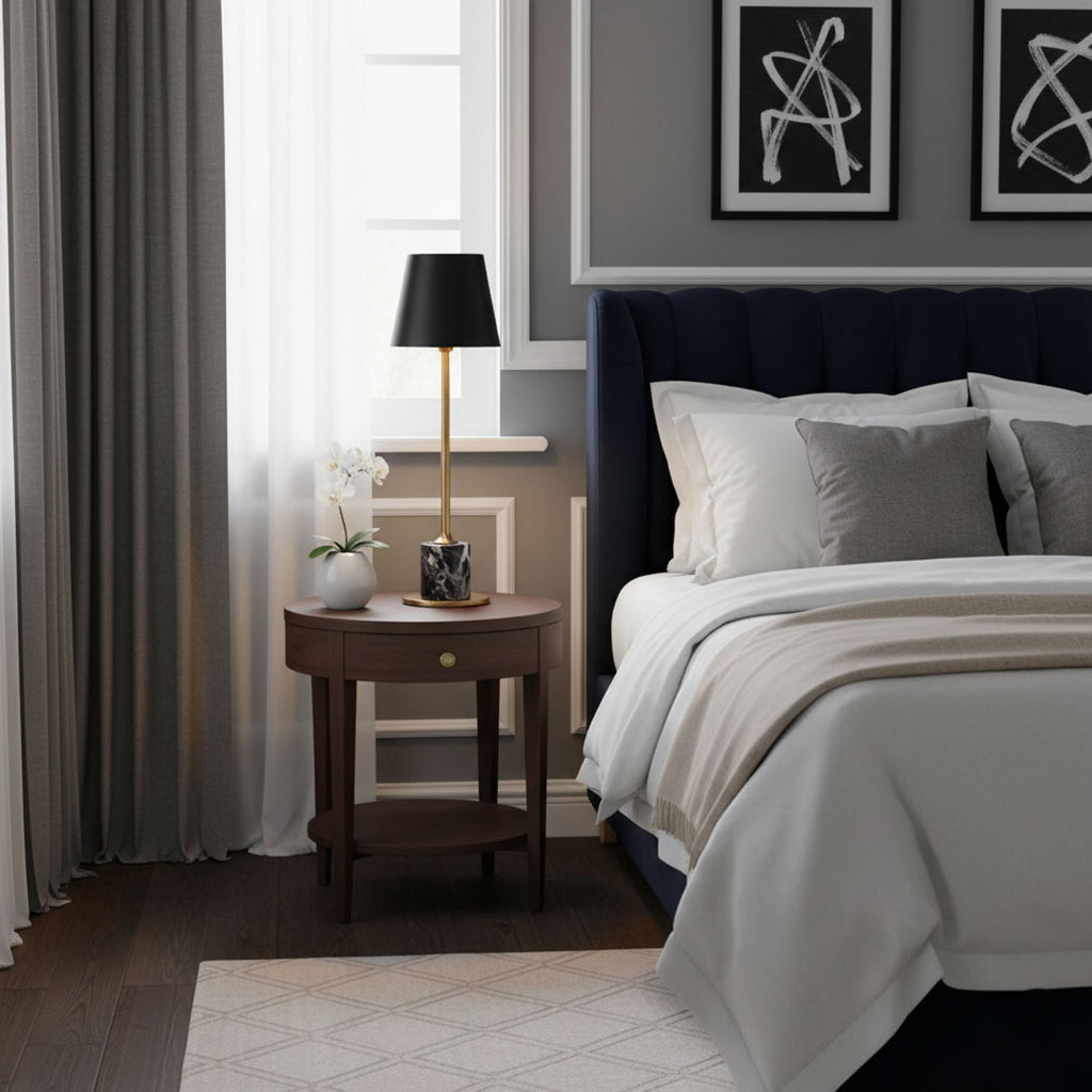 A Clérachic marble table lamp sits on a round nightstand beside a navy tufted bed, a small vase and sheer curtain nearby.