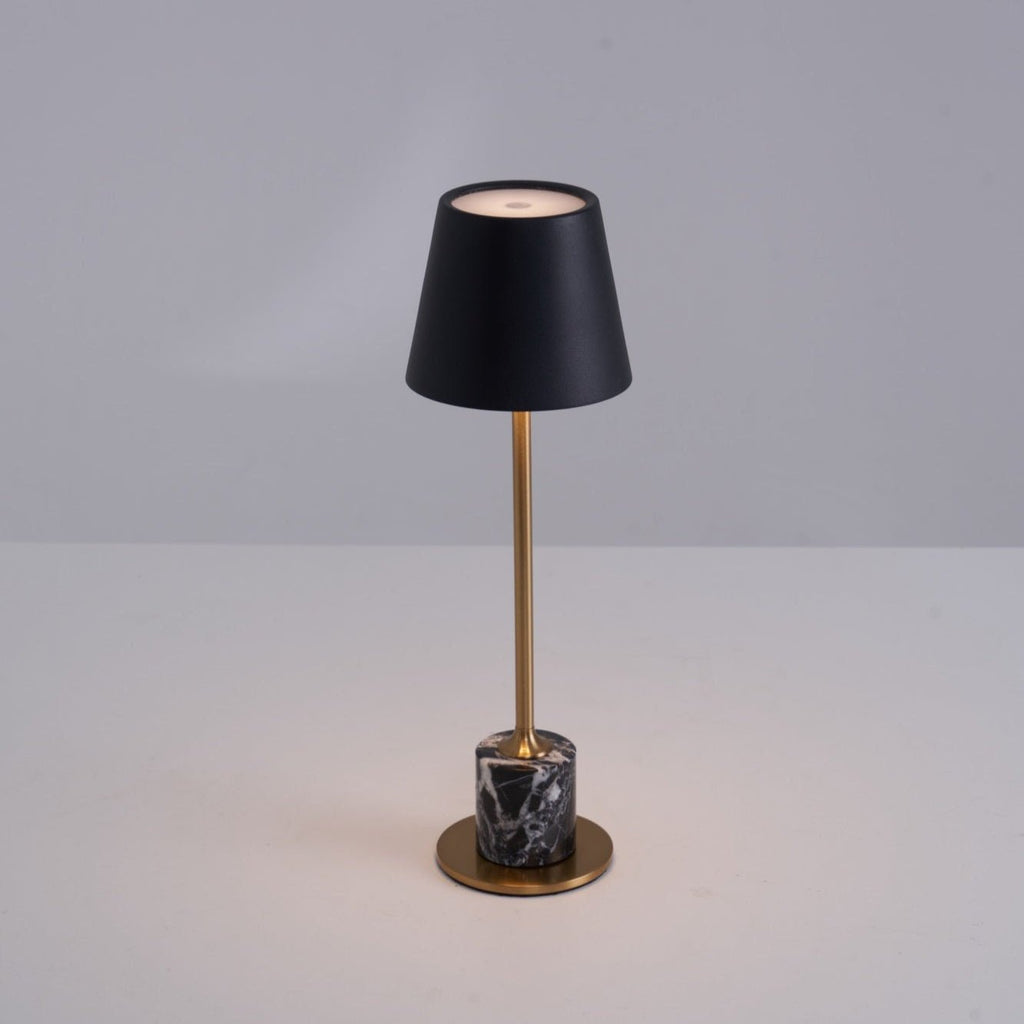 Lit warm on a plain tabletop, the black shade of this Clérachic lamp glows above its slim brass stem and dark marble cylinder base.