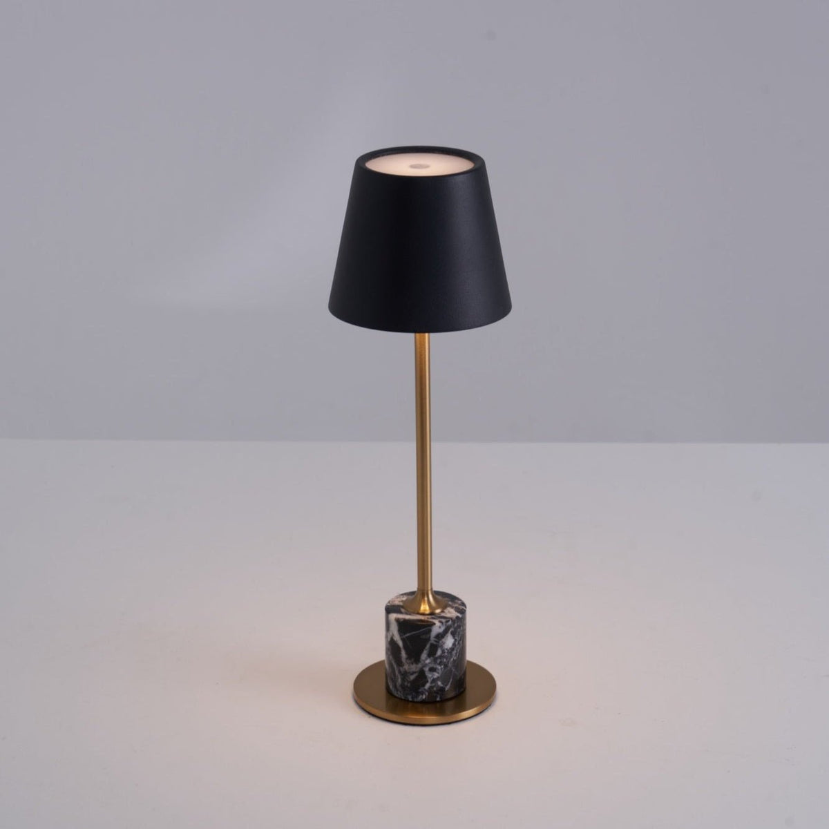 Lit warm on a plain tabletop, the black shade of this Clérachic lamp glows above its slim brass stem and dark marble cylinder base.