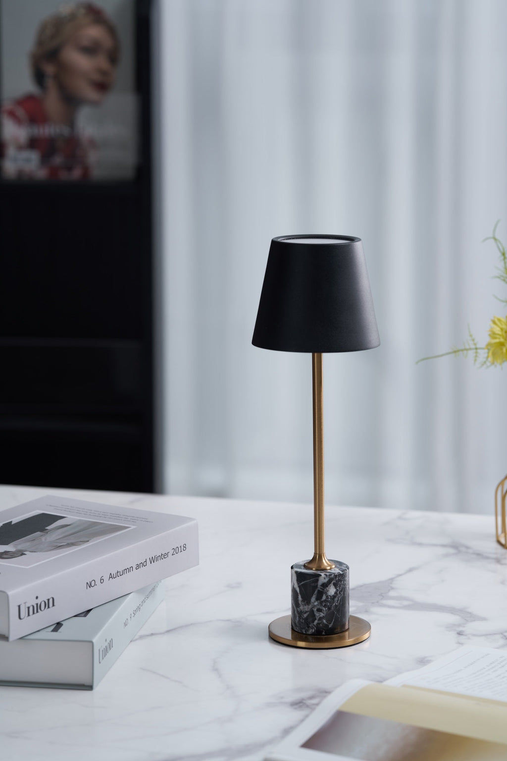 Set on a marble-topped nightstand beside stacked magazines, this Clérachic table lamp's black shade and brass stem stand against a softly blurred backdrop.