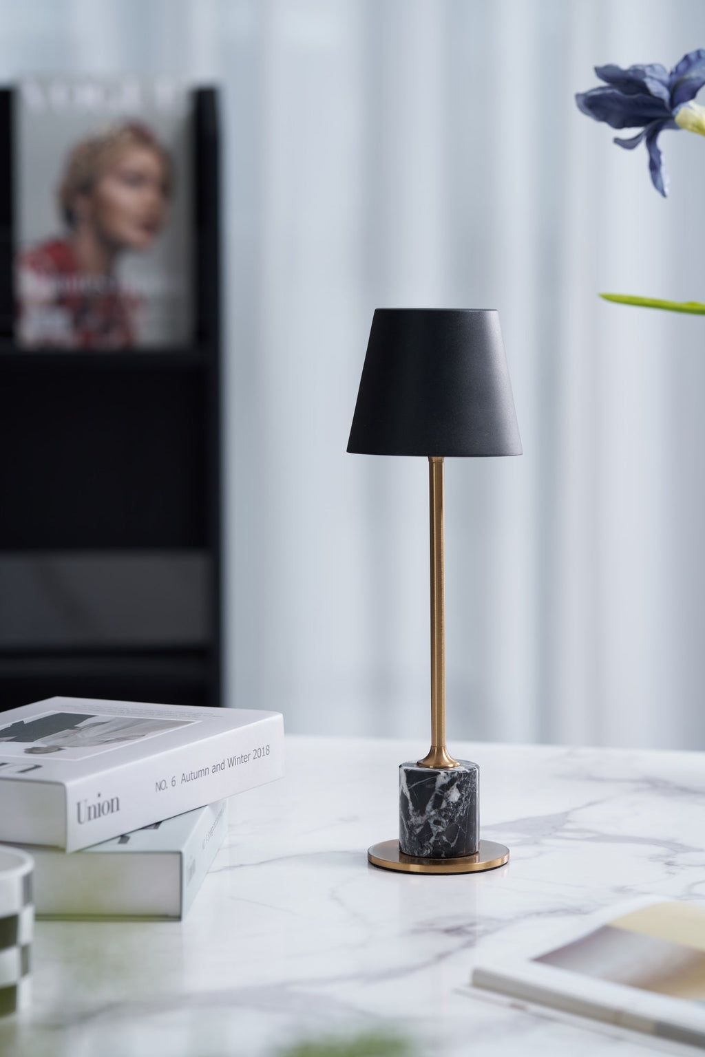 Styled beside a propped fashion magazine and a sprig of blue flowers, this Clérachic table lamp shows its black tapered shade on a marble-topped nightstand.