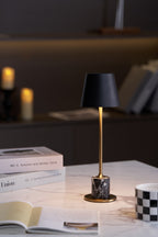 A Clérachic marble table lamp glows on marble beside a magazine stack and open book, a dark bookshelf blurred behind it.