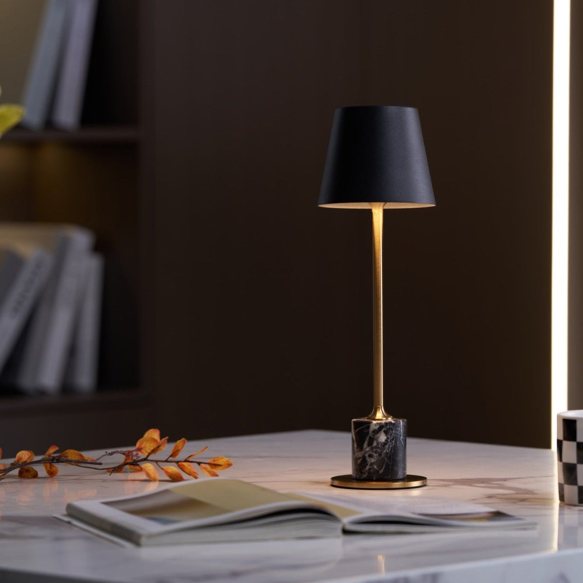 A Clérachic marble table lamp lights a marble tabletop beside an open magazine, a bookshelf blurred in the background.