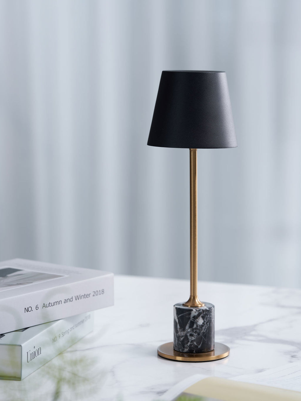 A Clérachic marble table lamp stands on a marble tabletop with a stacked magazine, a sheer curtain lit behind it.