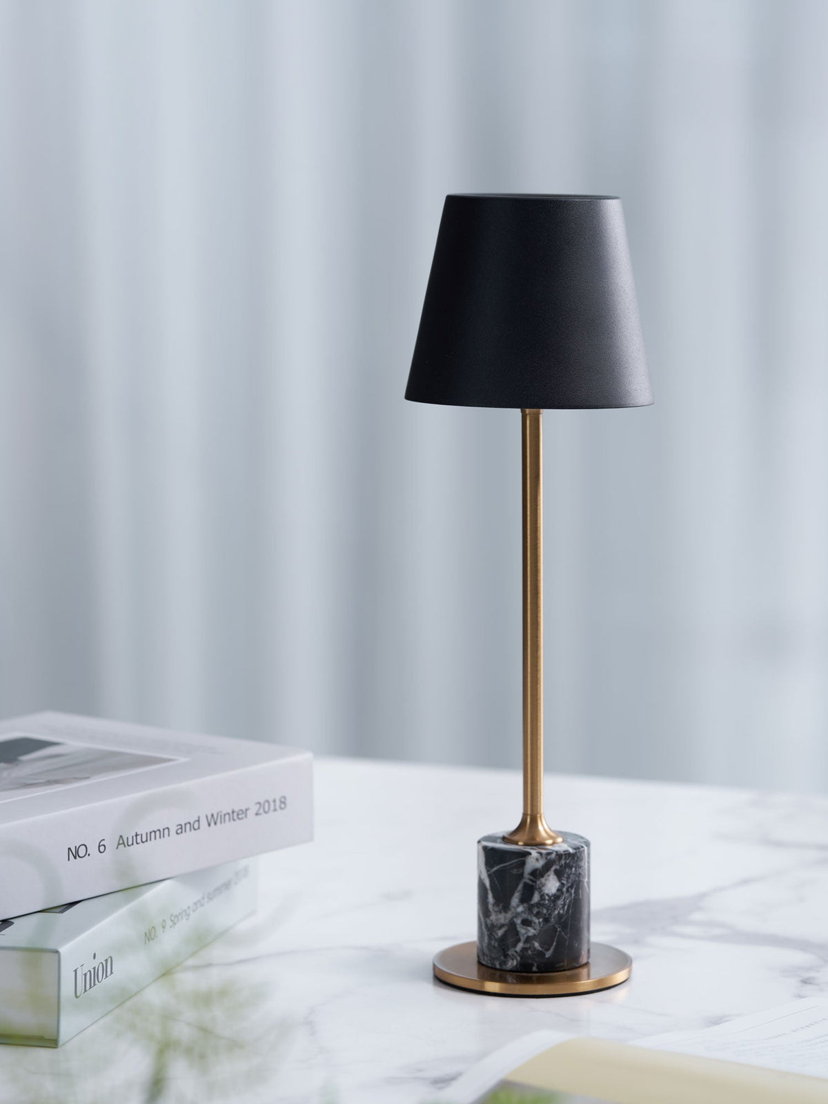 A Clérachic marble table lamp stands on a marble tabletop with a stacked magazine, a sheer curtain lit behind it.