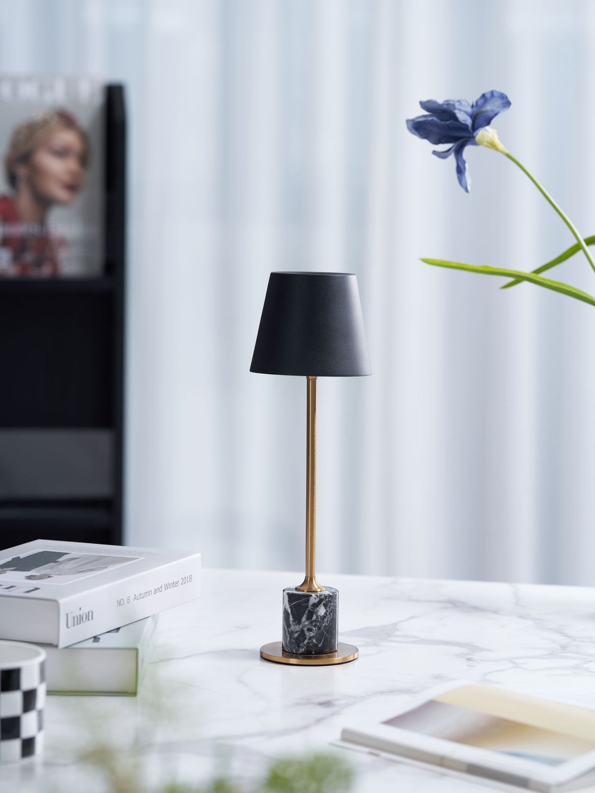 A Clérachic marble table lamp on a marble tabletop beside magazines, a blue flower stem placed just behind its shade.
