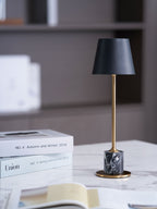A Clérachic marble table lamp glows on a marble tabletop beside a magazine stack, open book and mug, a bookshelf blurred behind.