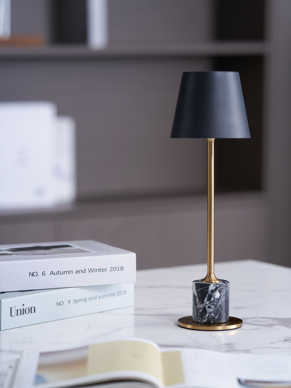 A Clérachic marble table lamp glows on a marble tabletop beside a magazine stack, open book and mug, a bookshelf blurred behind.