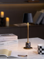 A Clérachic marble table lamp glows beside a magazine stack and open book, candlelight softly blurred in the background.