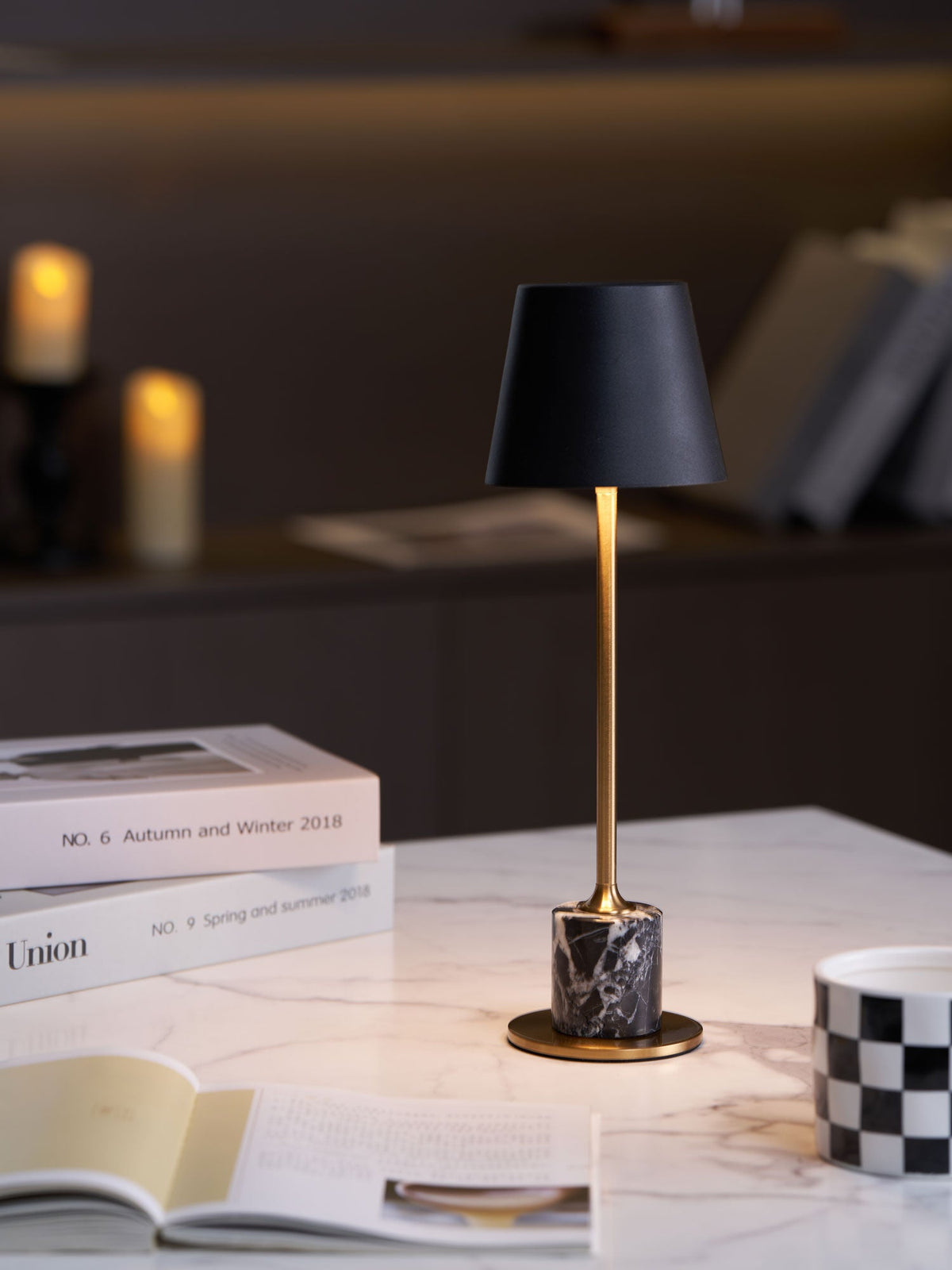 A Clérachic marble table lamp glows beside a magazine stack and open book, candlelight softly blurred in the background.