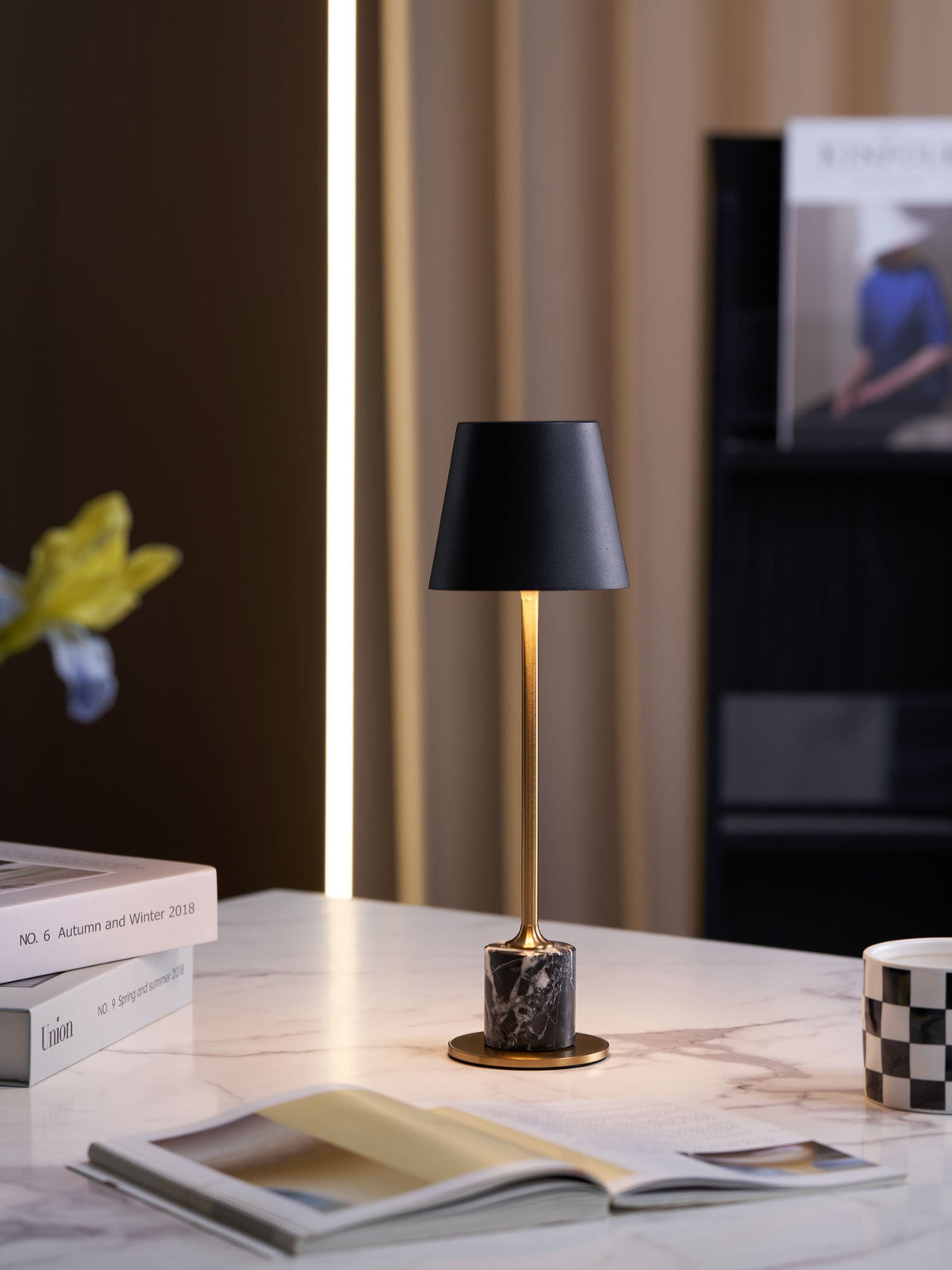 A Clérachic marble table lamp glows on marble with a dried orange branch, magazines and a chequered mug arranged around its base.