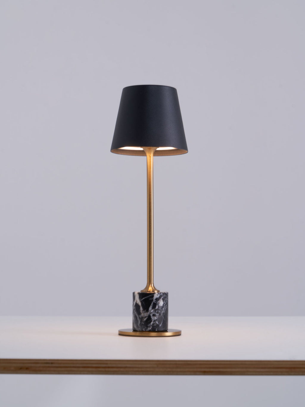 Set at the edge of a wooden tabletop, this Clérachic table lamp's black shade glows warm above its brass stem and dark marble base.