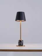 Set at the edge of a wooden tabletop, this Clérachic table lamp's black shade glows warm above its brass stem and dark marble base.