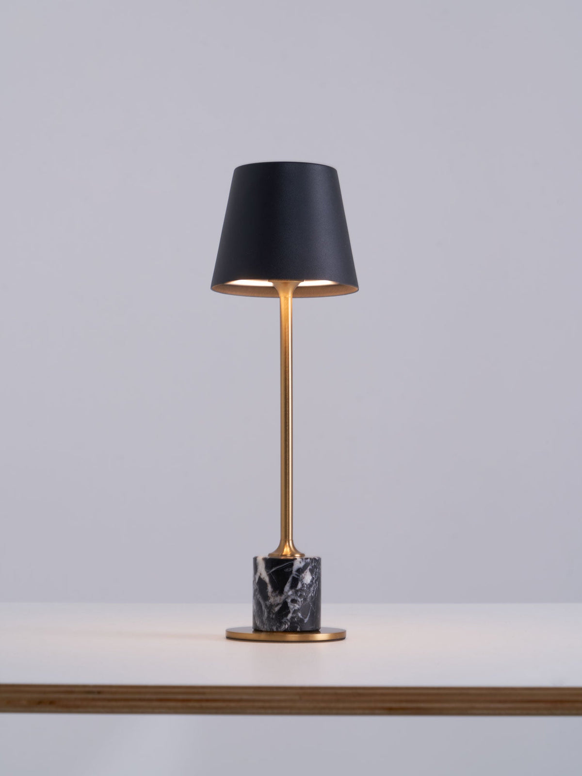 Set at the edge of a wooden tabletop, this Clérachic table lamp's black shade glows warm above its brass stem and dark marble base.