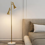 A beige bed with white pillows sits beside a brass Clérachic floor lamp glowing warm on its white marble base.