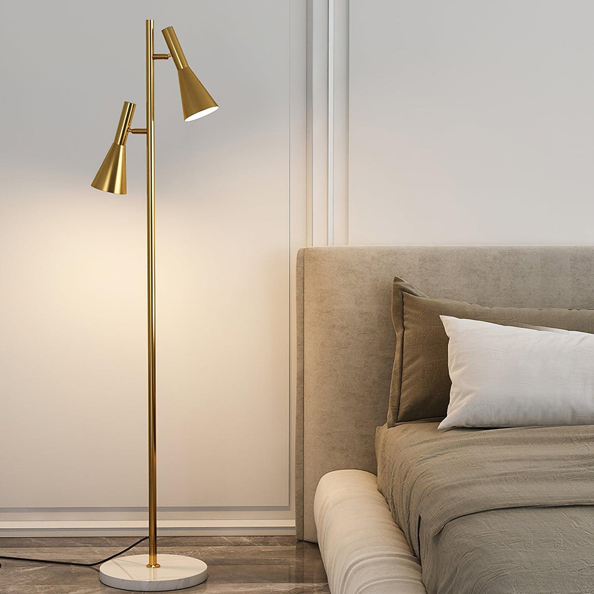 A beige bed with white pillows sits beside a brass Clérachic floor lamp glowing warm on its white marble base.
