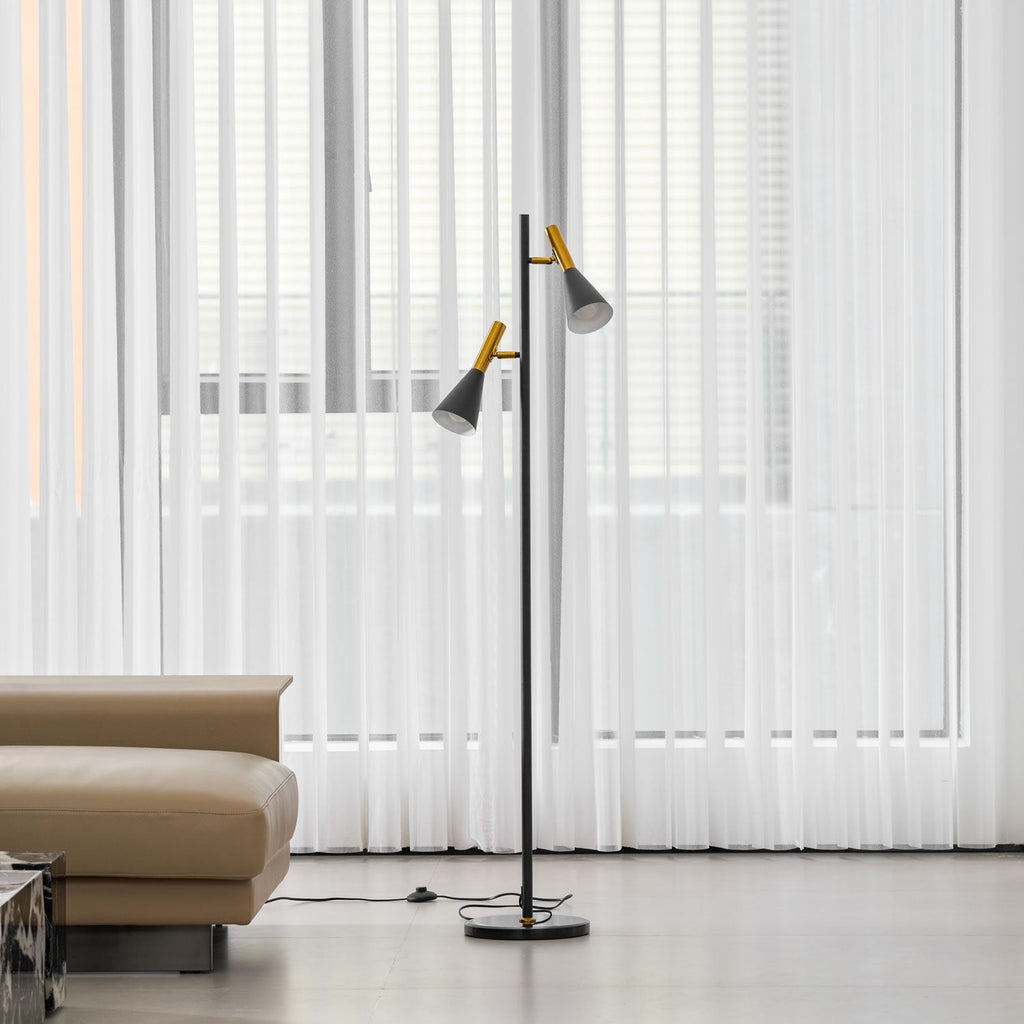 Black Clérachic floor lamp with brass-necked shades standing between sheer curtains and a beige leather sofa.