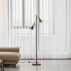 Black Clérachic floor lamp with brass-necked shades standing between sheer curtains and a beige leather sofa.