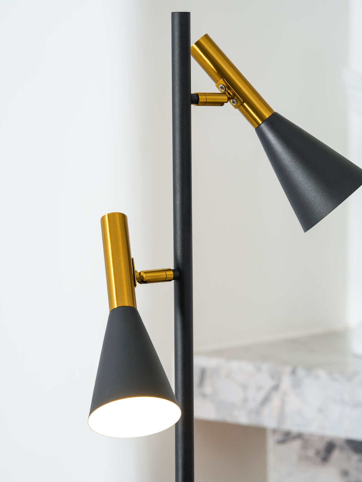 Close-up of a brass neck and black cone shade on a Clérachic floor lamp, a marble mantel blurred behind it.